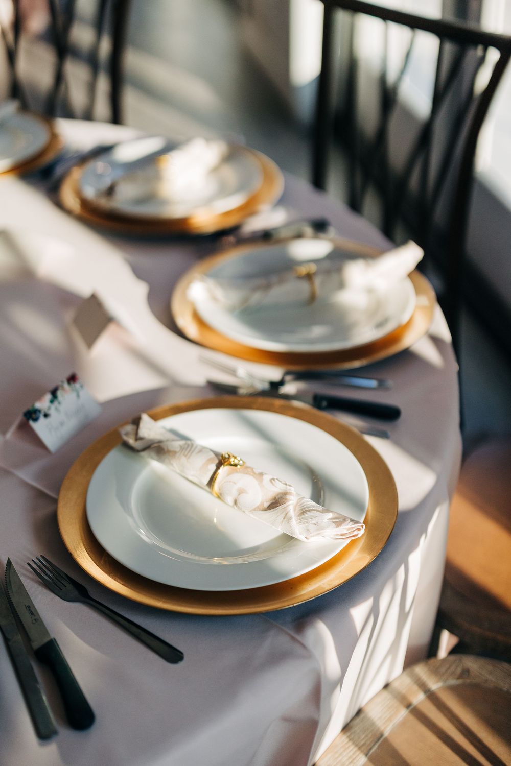 Elegant table settings with white plates, gold chargers, and dark cutlery arranged for a formal dining event.