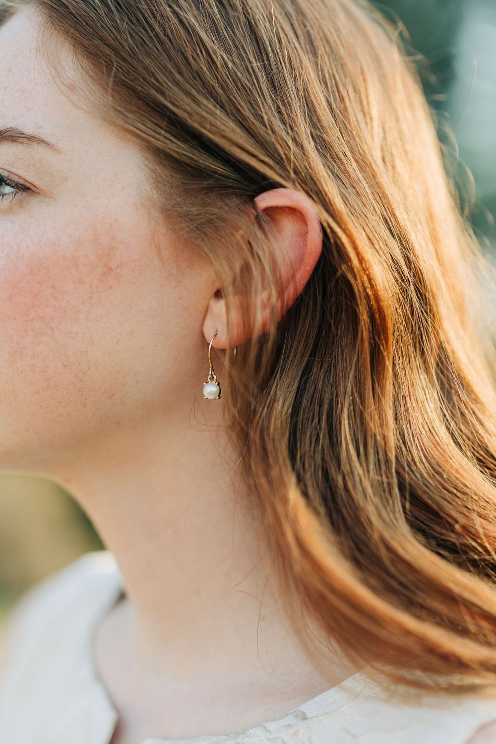 Close-up profile view of someone with wavy brown hair wearing delicate drop earrings in soft natural lighting.