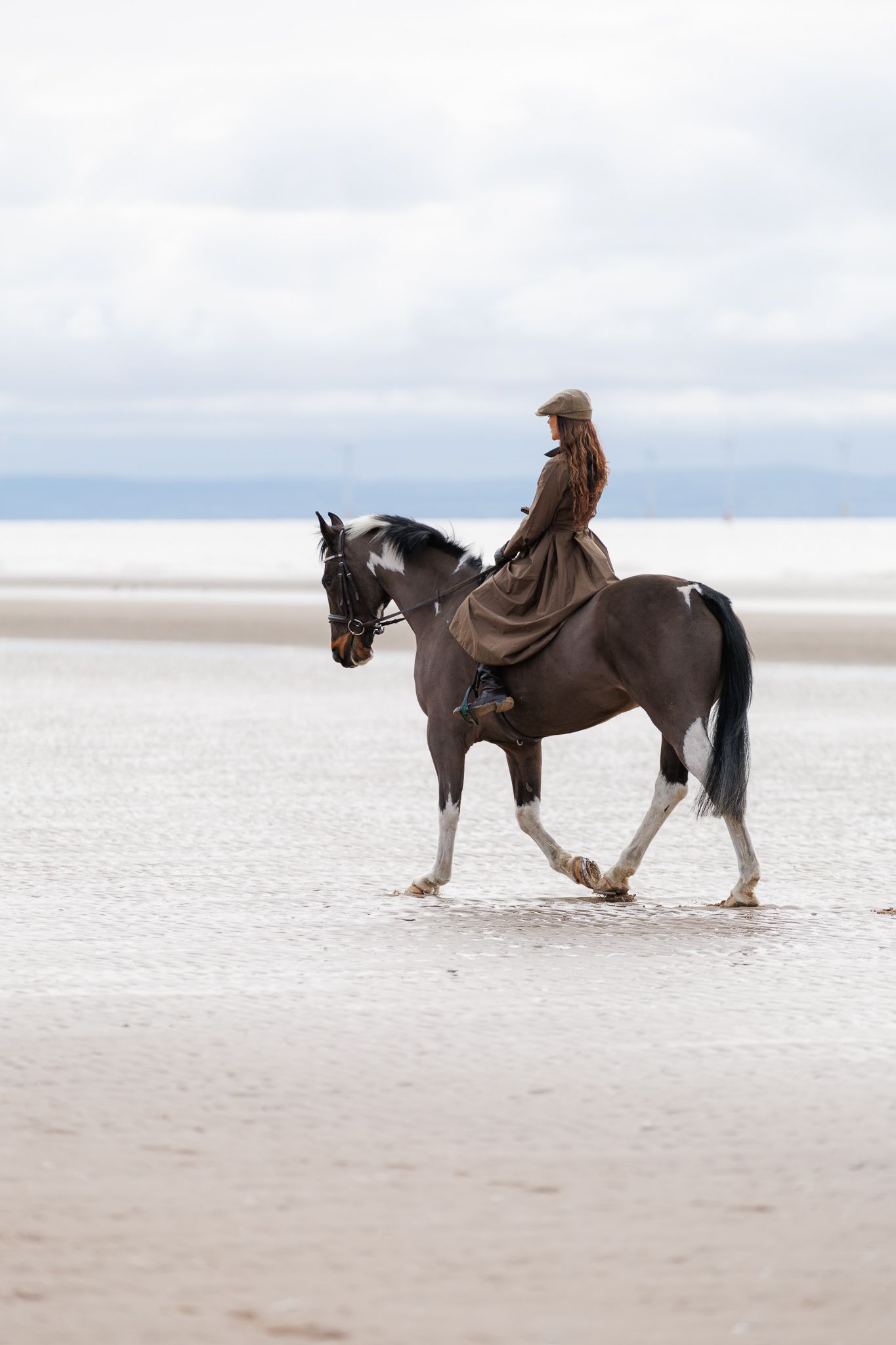 A rider in brown clothing guides a black and white horse along a serene, misty beach shoreline.