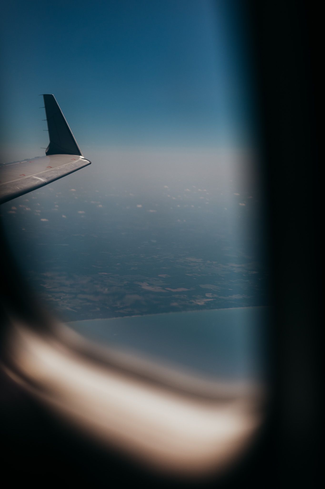 View from airplane window showing wing silhouette against blue sky and distant landscape below.