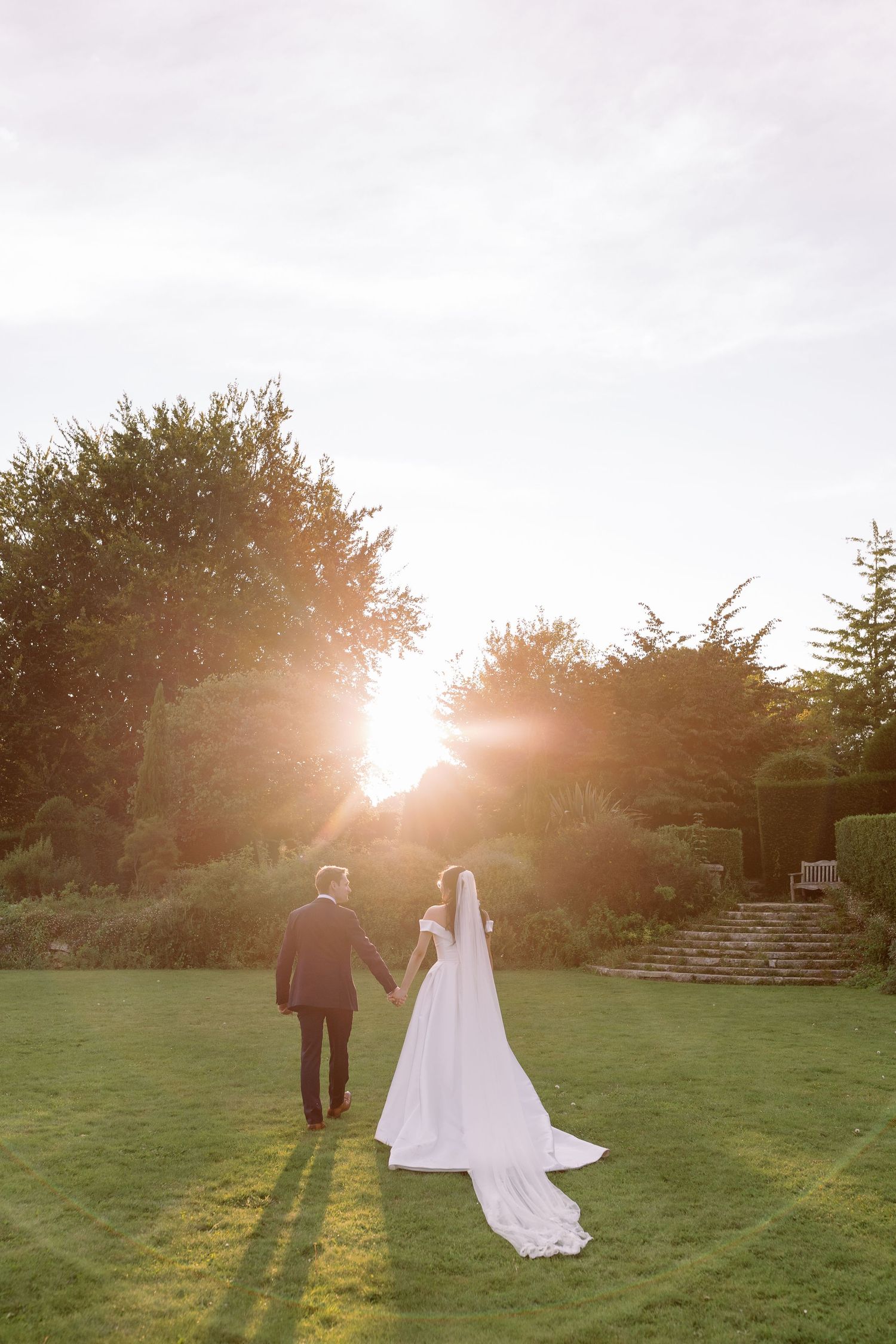 A bride and groom walk across a sunlit lawn at golden hour, creating long shadows against the grass at Kingston Maurward by Maddie Waters