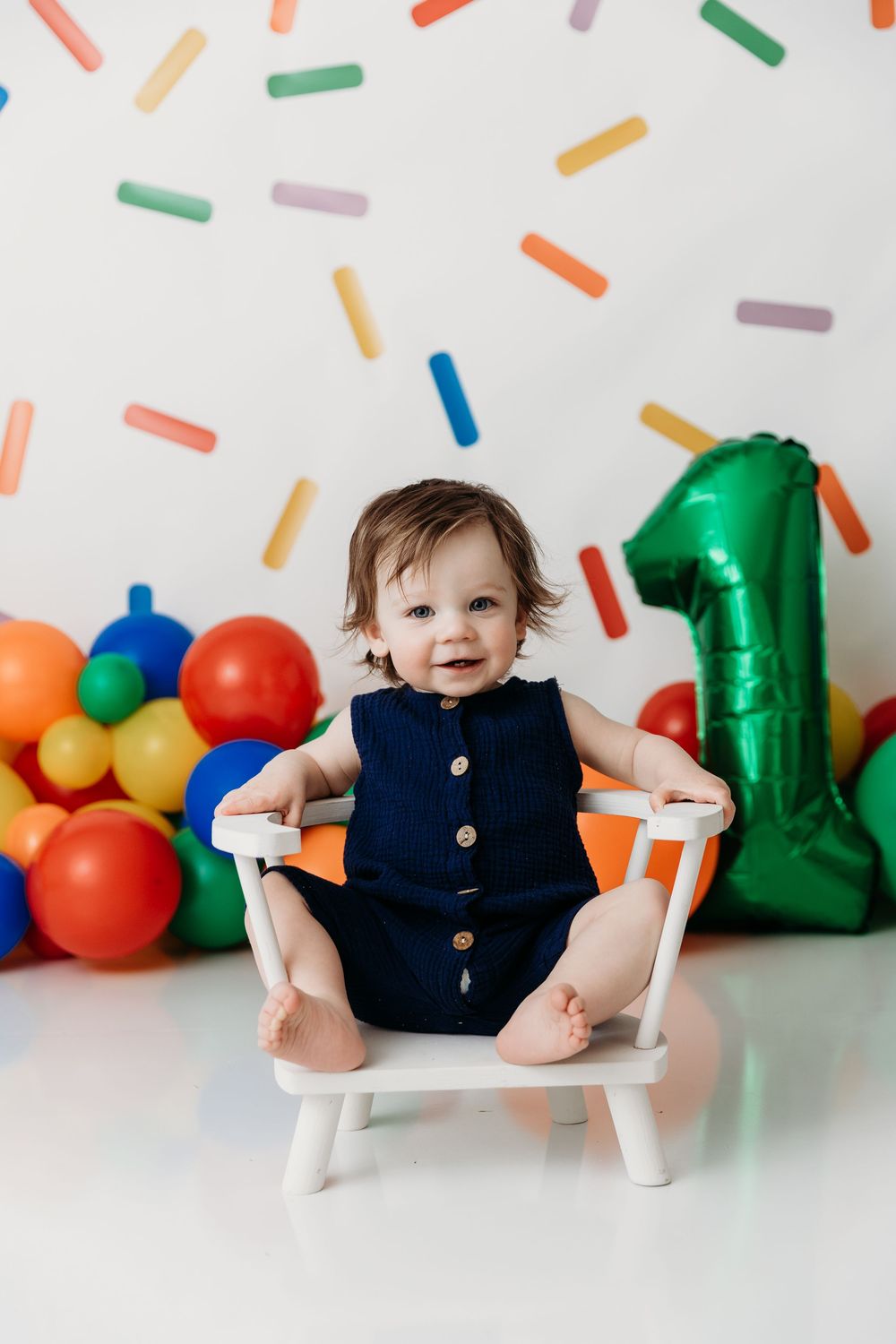 Small child in navy dress sits in white chair against colorful balloon and sprinkle backdrop for cake smash photo.