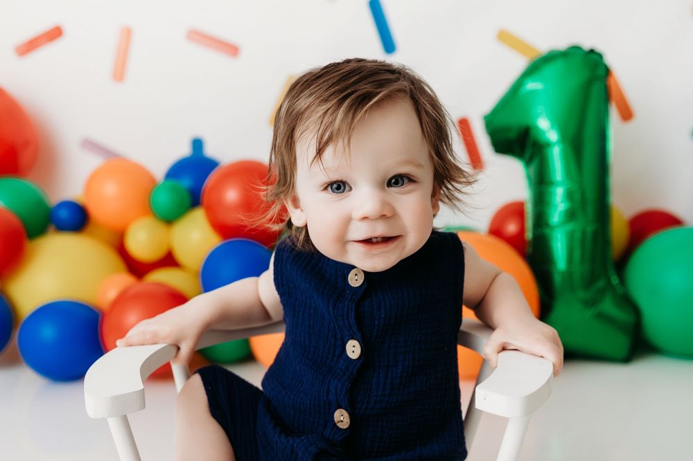 First birthday party setup with colorful balloons and a navy blue outfit against a white backdrop.