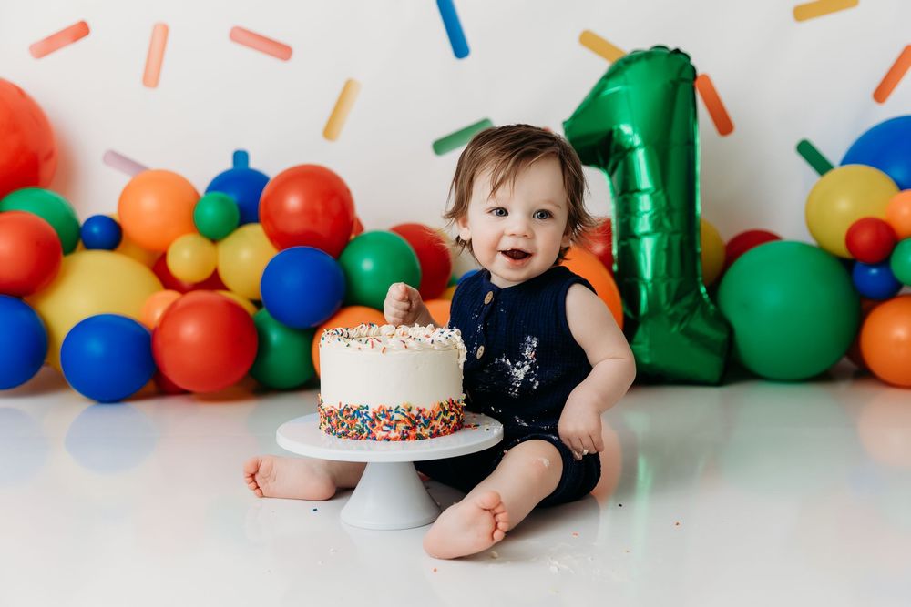 A festive first birthday party setup with colorful balloons and cake smash photos against a white backdrop.