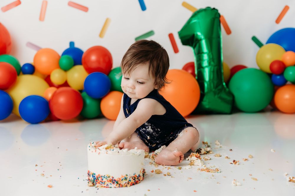 A child smashes their face into a white frosted cake with colorful sprinkles while surrounded by balloons.