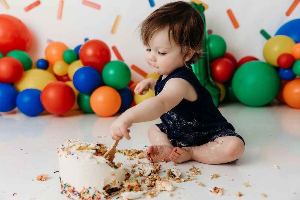 A baby sits on the floor surrounded by colorful balloons while enjoying a slice of birthday cake.