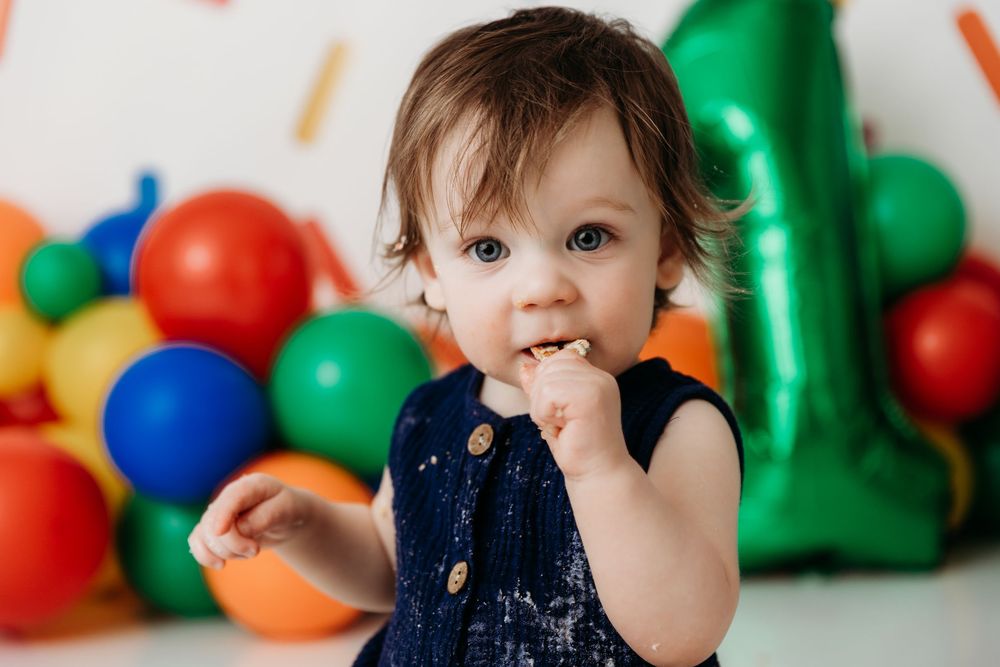 A toddler in a navy dress smiles while playing with colorful balloons at a first birthday party celebration.