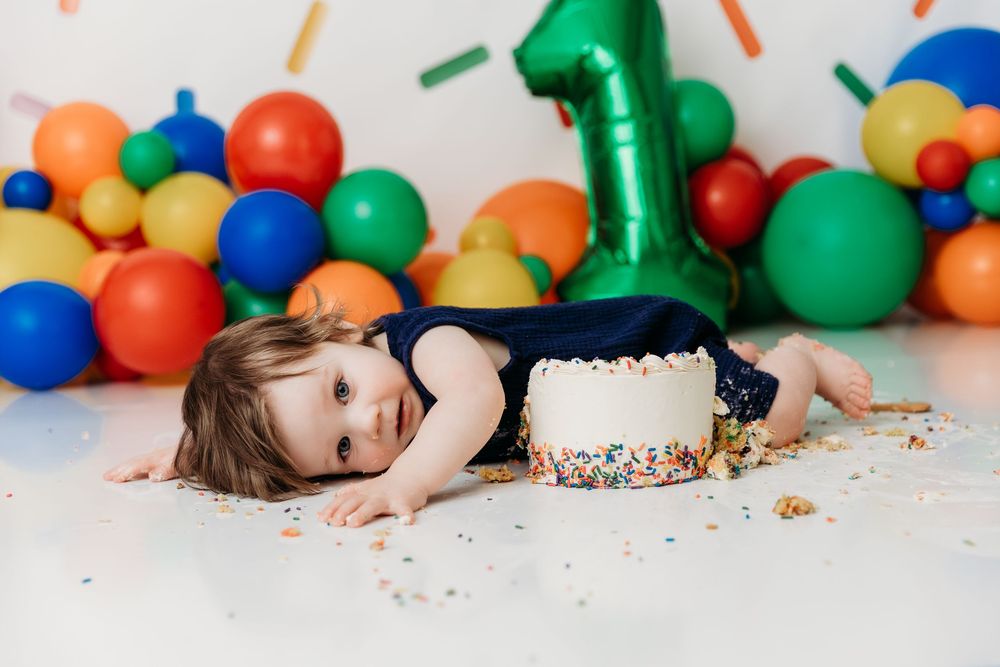 Colorful first birthday cake smash photo session with balloons and scattered sprinkles on white backdrop.