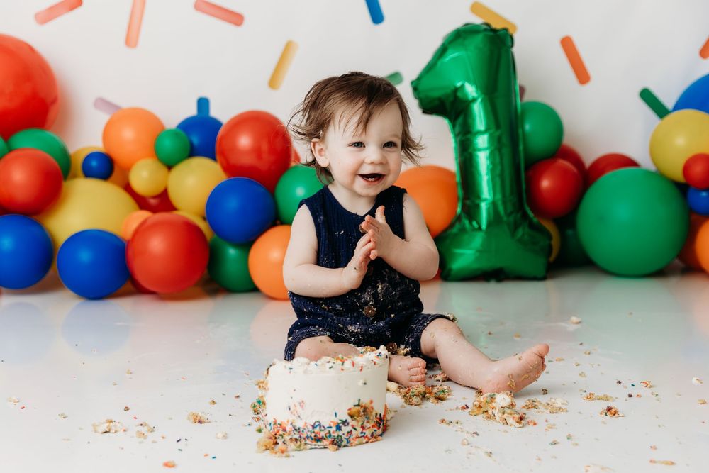 Colorful cake smash photo session with balloons and decorations as a baby explores their first birthday cake.