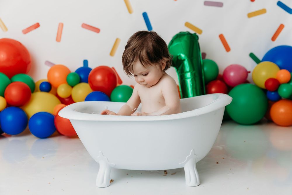 Baby taking bath in white tub surrounded by colorful balloons against white background.
