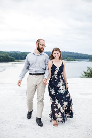 A couple walks hand in hand along a white sandy beach with a lake and trees in the background.
