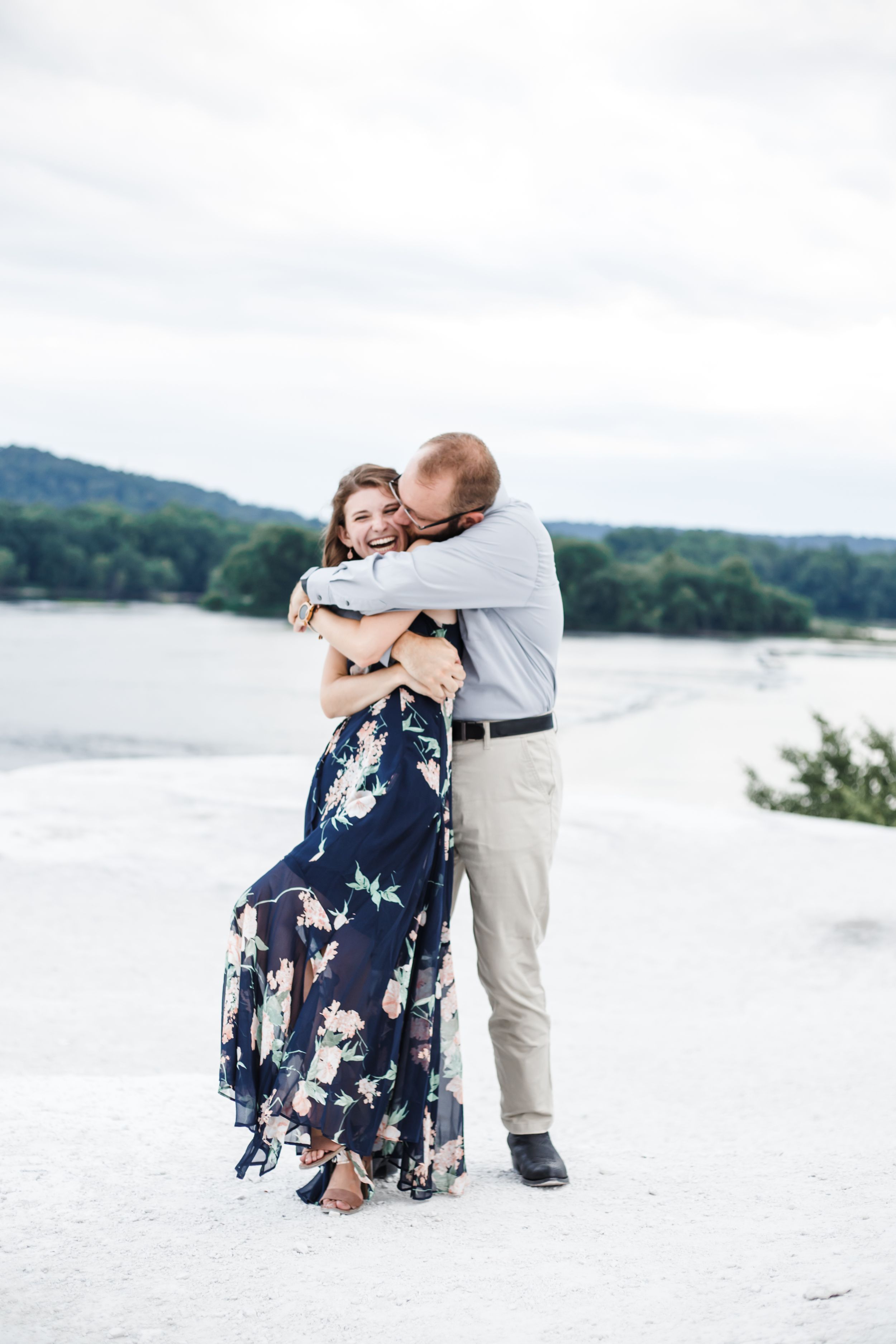 A couple dances together on a white sandy beach with trees and water in the background.