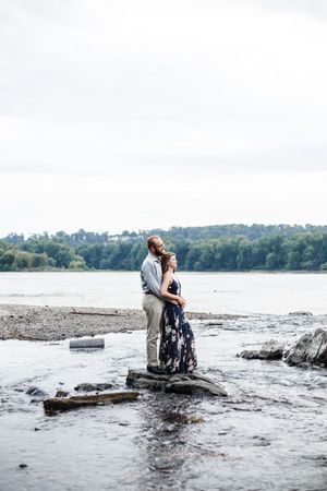 Couple embracing on rocky shoreline with lake and forest backdrop during engagement photoshoot.