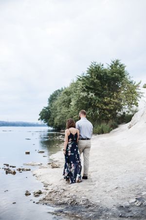 A couple walks along a scenic rocky shoreline with trees and water in the background.