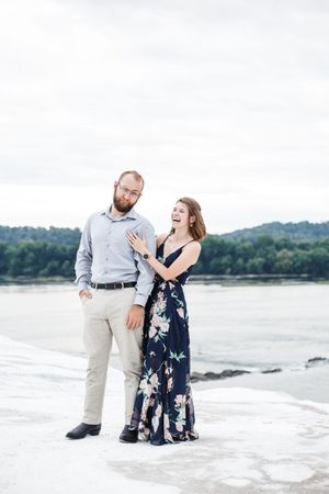 A couple shares a joyful moment together on a rocky shoreline with mountains and water in the background.