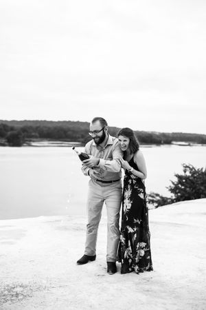A couple laughs together while sitting on a white rocky outcrop with a serene lake landscape in the background.