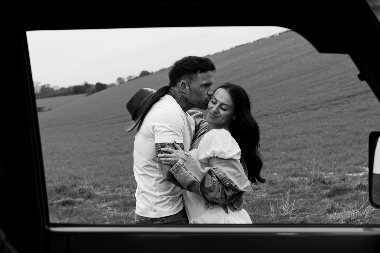 Couple sharing a tender kiss framed by a car window with open fields in the background.