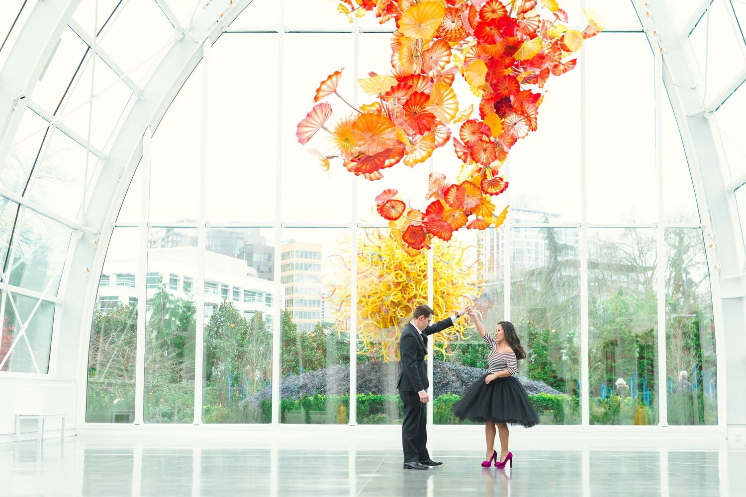 Couple poses beneath vibrant red and orange Chihuly glass sculpture in a bright modern gallery with floor-to-ceiling windows.