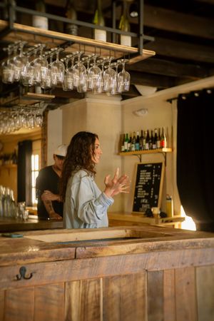 Woman bartender gesturing behind a rustic wooden bar with hanging wine glasses and bottle display.