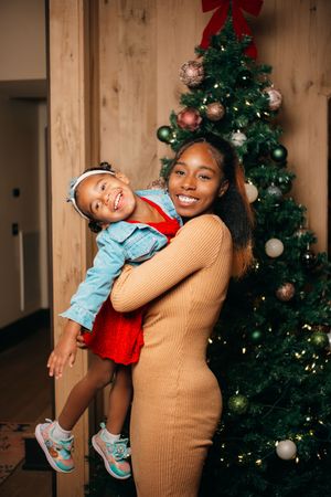 A woman and young girl smile together in front of a decorated Christmas tree during the holiday season.