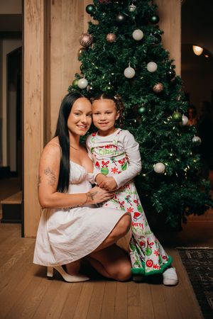 A woman in a white dress kneels beside a young girl in Christmas pajamas in front of a decorated Christmas tree.
