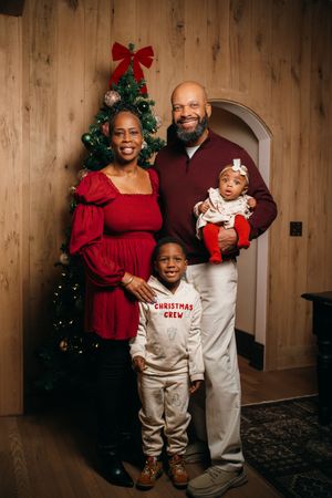 Family of four poses by Christmas tree, parents in red and maroon holding baby, son wearing Christmas Crew outfit.