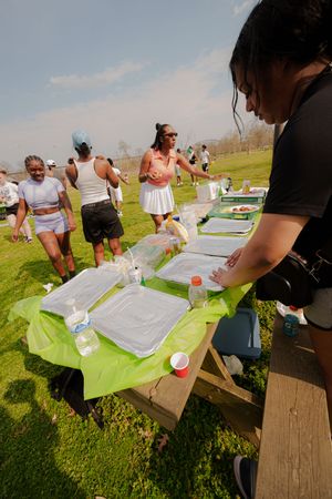 People gathered outdoors at a picnic table with trays and supplies during a sunny day event in a green park.