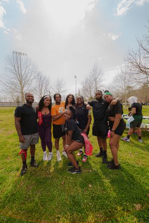 A group of nine friends posing together outdoors in a park on a cloudy day, smiling and wearing athletic clothing.