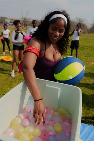Woman in purple top holding beach ball reaches into bin of colorful water balloons outdoors.