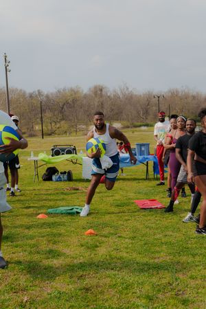 Athletes training outdoors on grass field with cones, a man carrying a ball during a football drill with spectators watching.