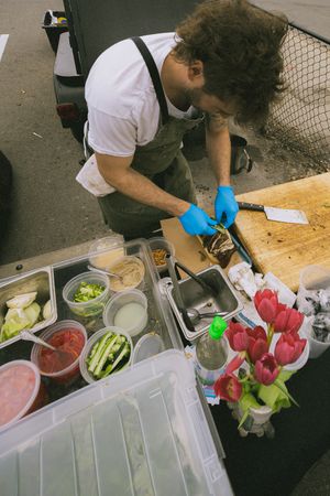 A curly-haired chef in blue gloves prepares food at an outdoor market stall with fresh ingredients and red tulips.