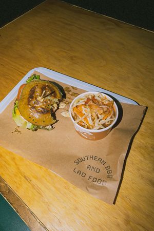 Southern BBQ and Lao food tray with a burger and a cup of coleslaw on a wooden table.