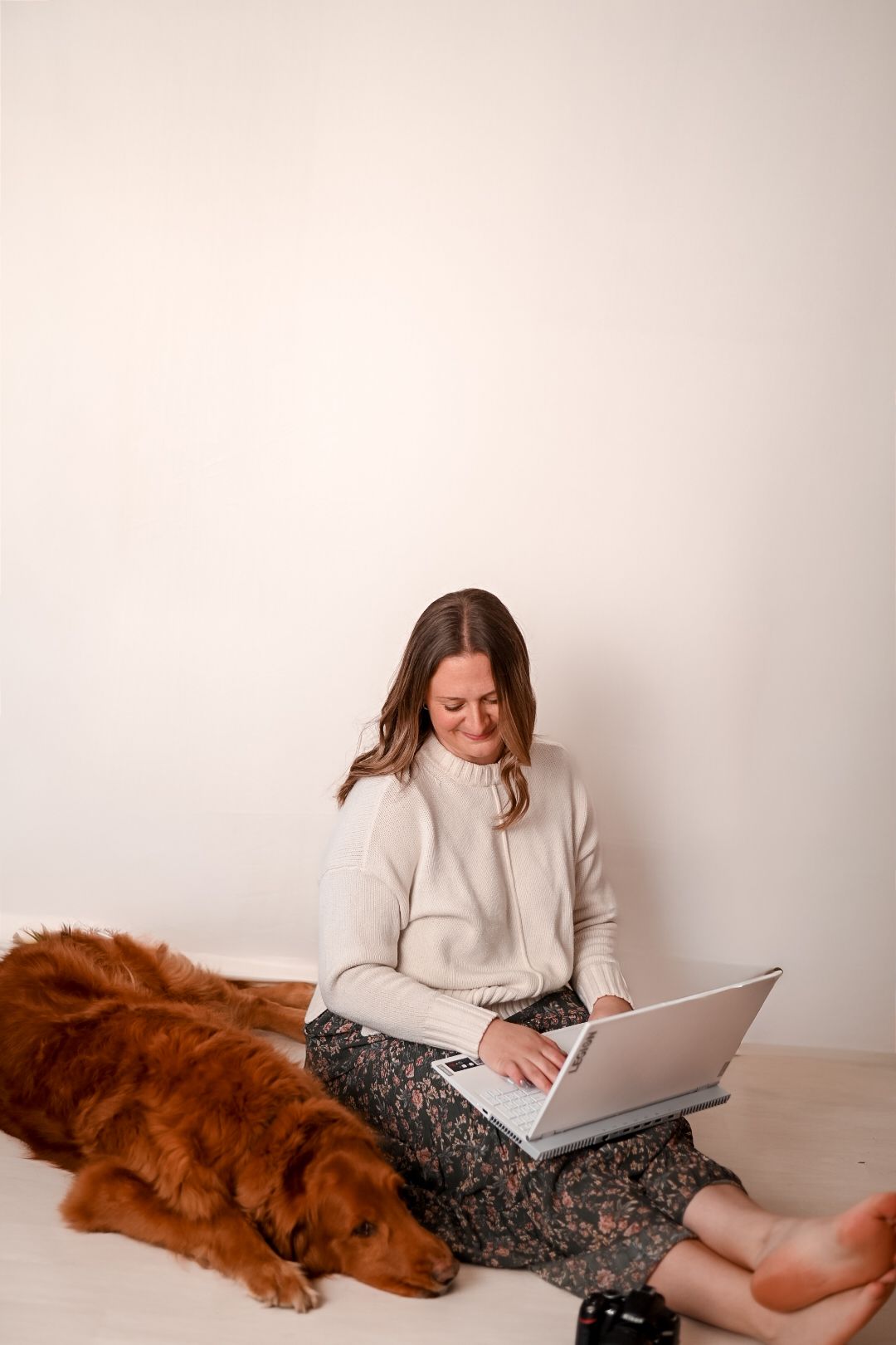 Woman in cream sweater sits cross-legged on floor working on laptop beside a fluffy red Golden Retriever dog.