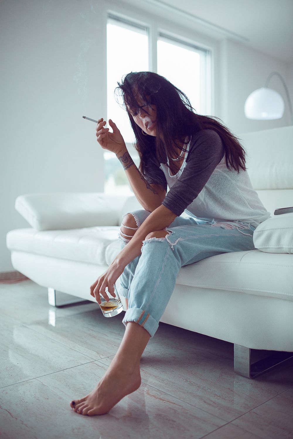 Porträtaufnahme einer tätowierten Frau, die rauchend mit einem Glas Drink auf einem weissen Sofa sitzt und nachdenklich nach unten blickt.