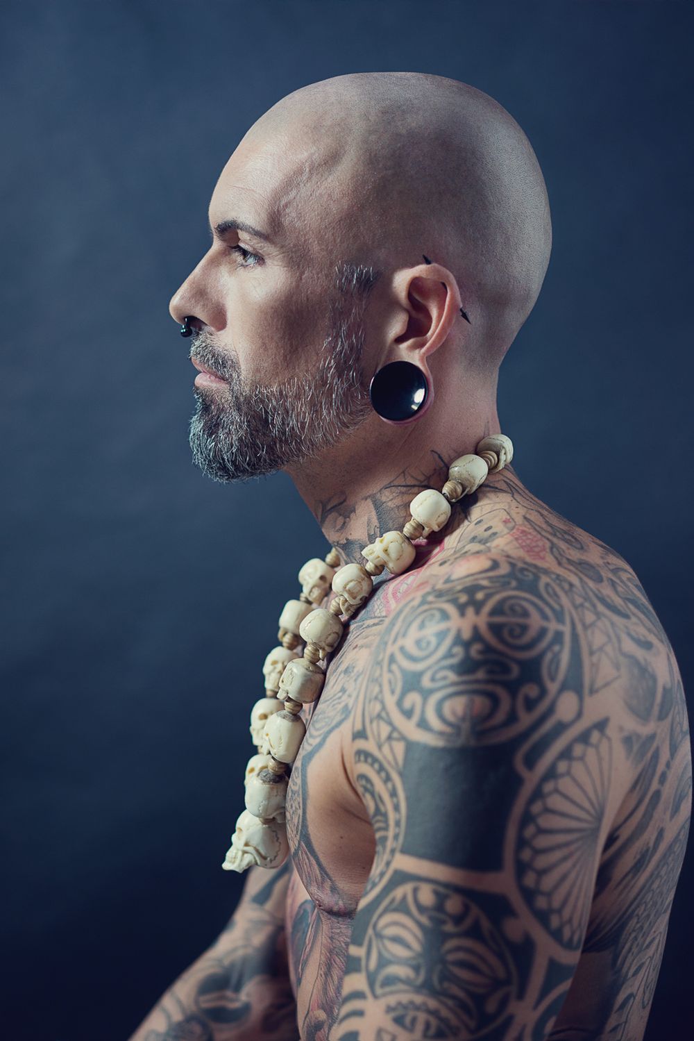 Seitenprofil eines stark tätowierten, kahlköpfigen Mannes mit Bart, Piercings und einer Kette aus Totenköpfen vor dunklem Hintergrund.