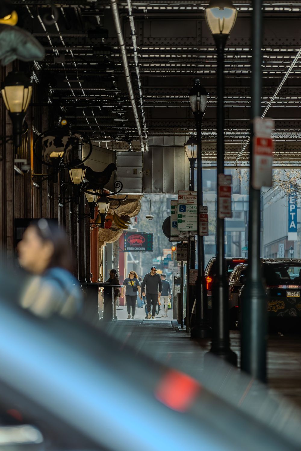 People walking under an elevated urban structure lined with vintage street lamps in a city neighborhood.