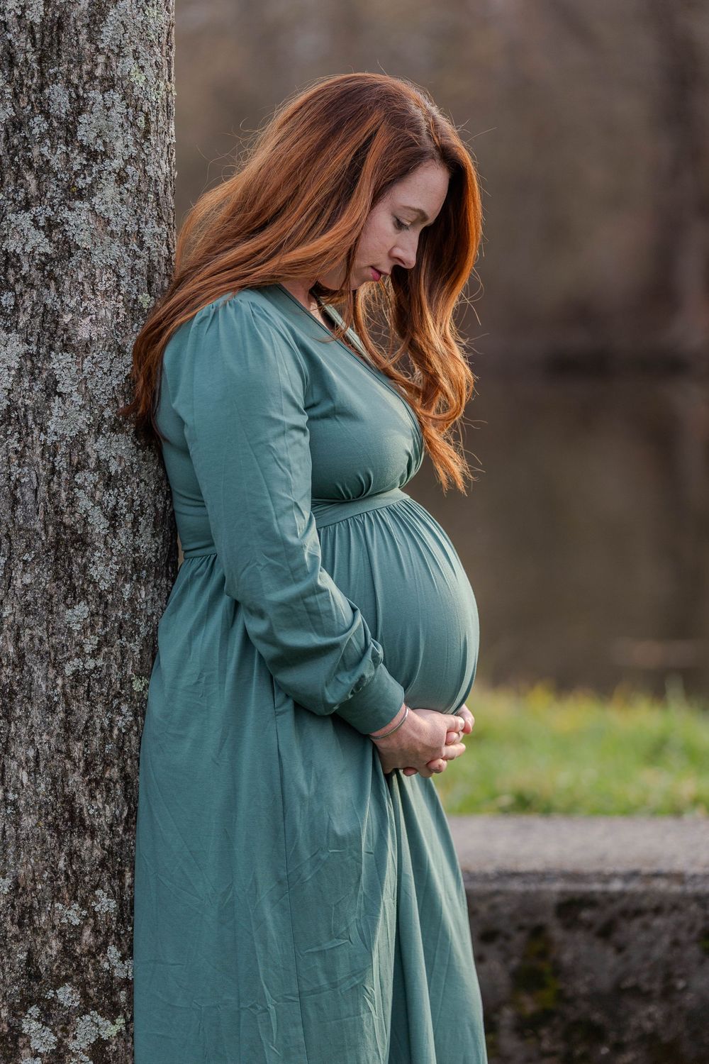 A pregnant woman in a teal dress stands thoughtfully by a stone wall in a natural outdoor setting.