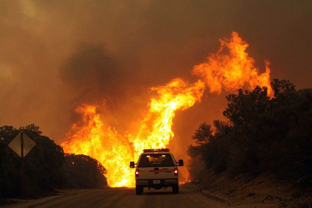 Intense wildfire rages through a dark landscape with flames engulfing vehicles and trees against a smoky night sky.