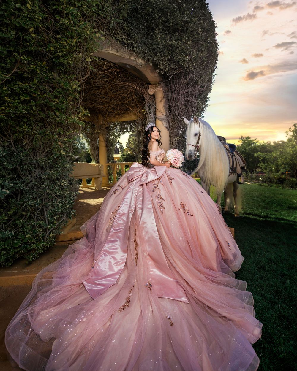 A flowing pink ball gown with intricate details photographed in a garden setting during golden hour sunset.