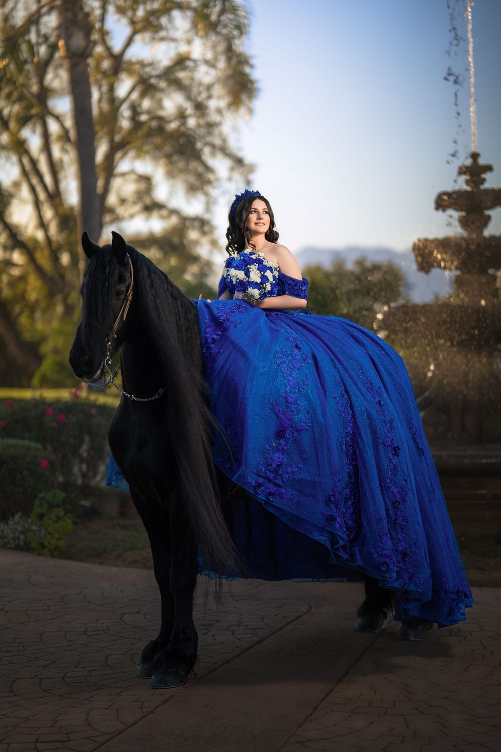 A person in a flowing royal blue ballgown poses elegantly with a black horse in an outdoor garden setting at sunset.