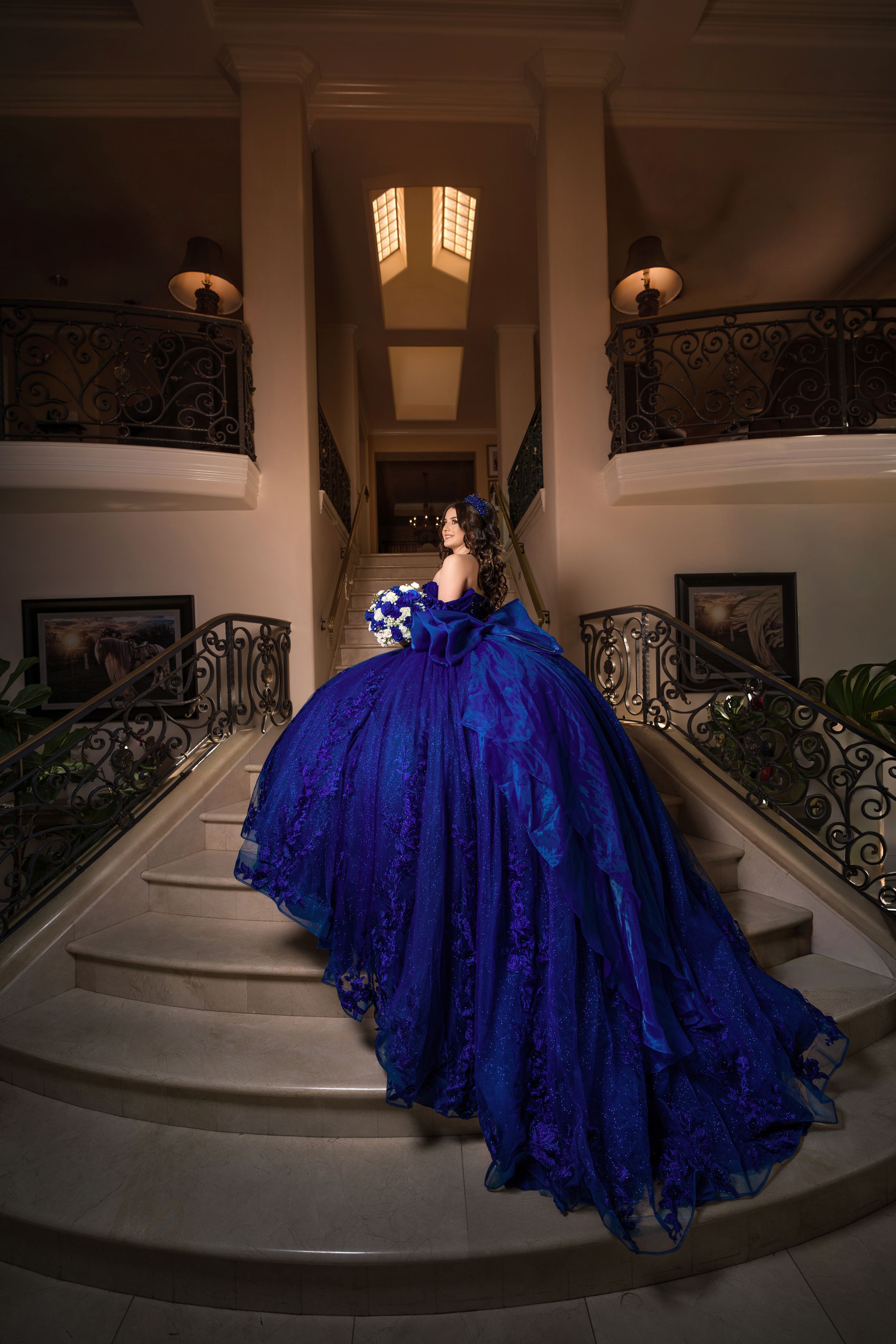A stunning royal blue ballgown with intricate lace details cascades down an elegant staircase with ornate iron railings.
