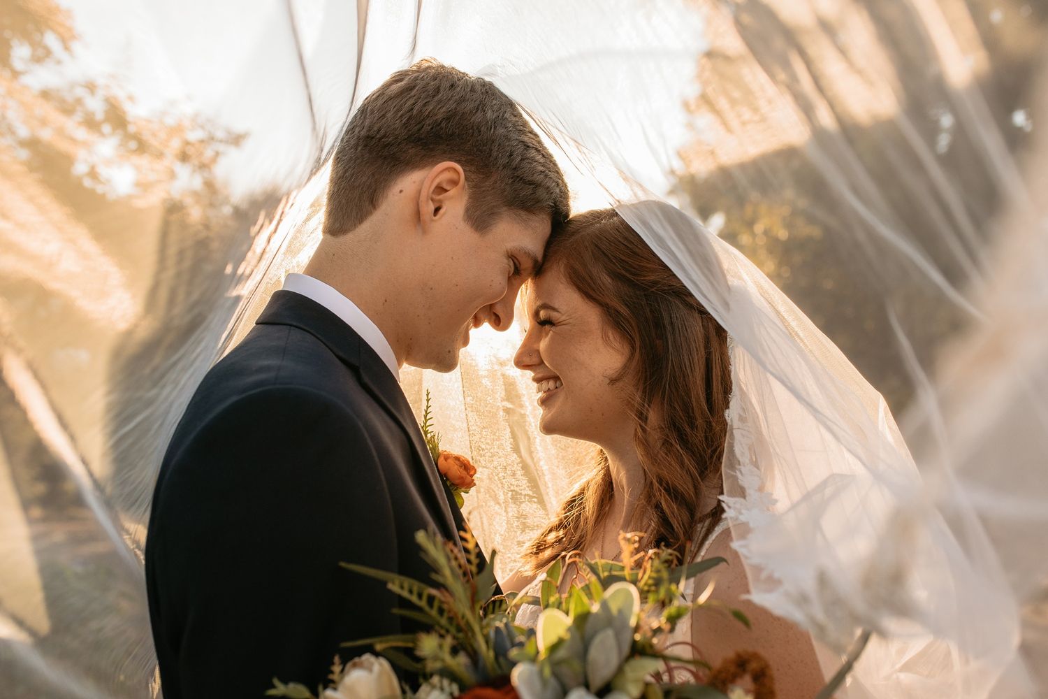 Bride and groom share a romantic moment under a wedding veil, with soft golden sunlight and a floral bouquet at Rancho Buena Vista Adobe.