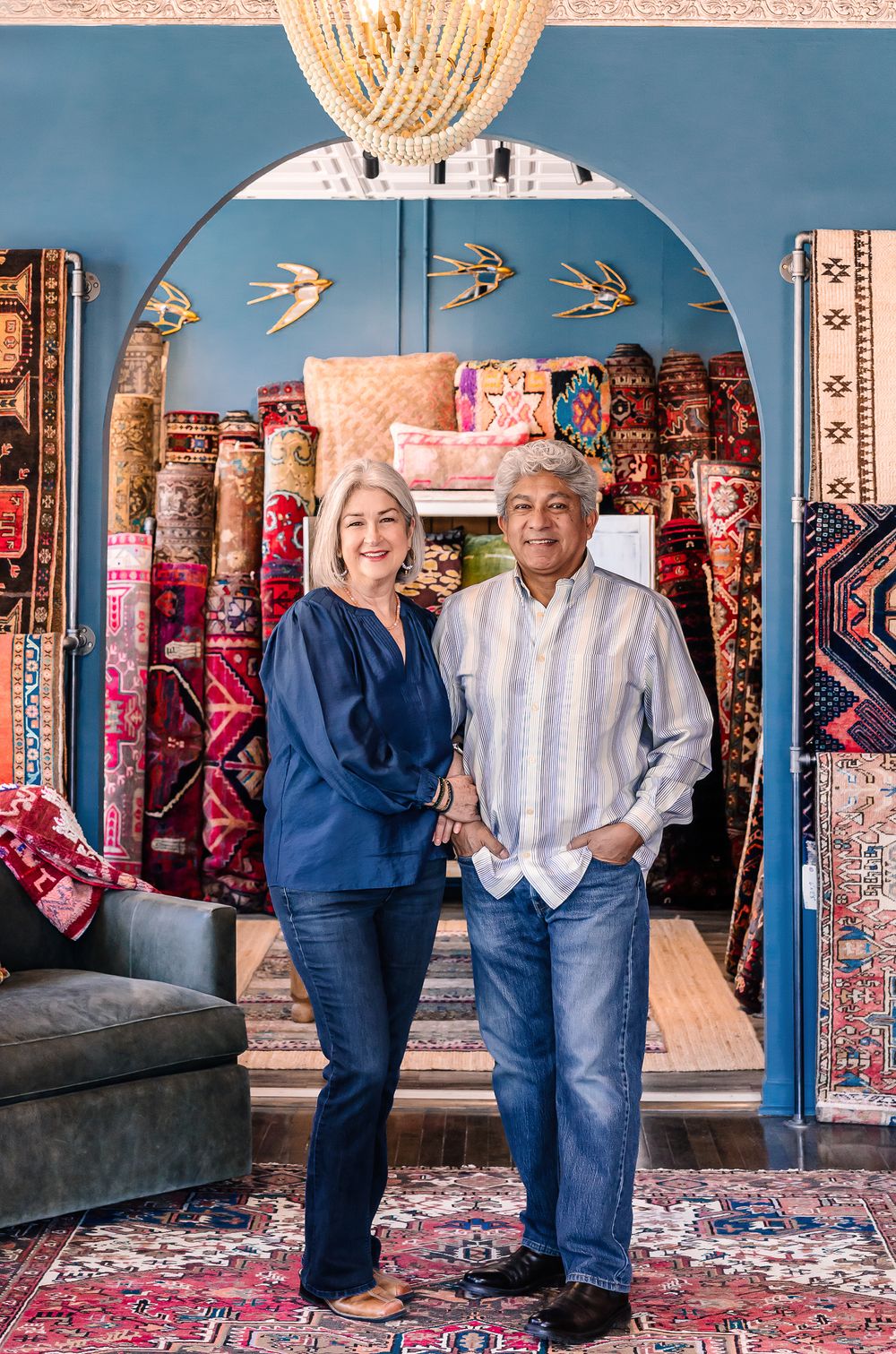 Couple smiling in a colorful rug shop with stacked Persian rugs and blue arched walls.