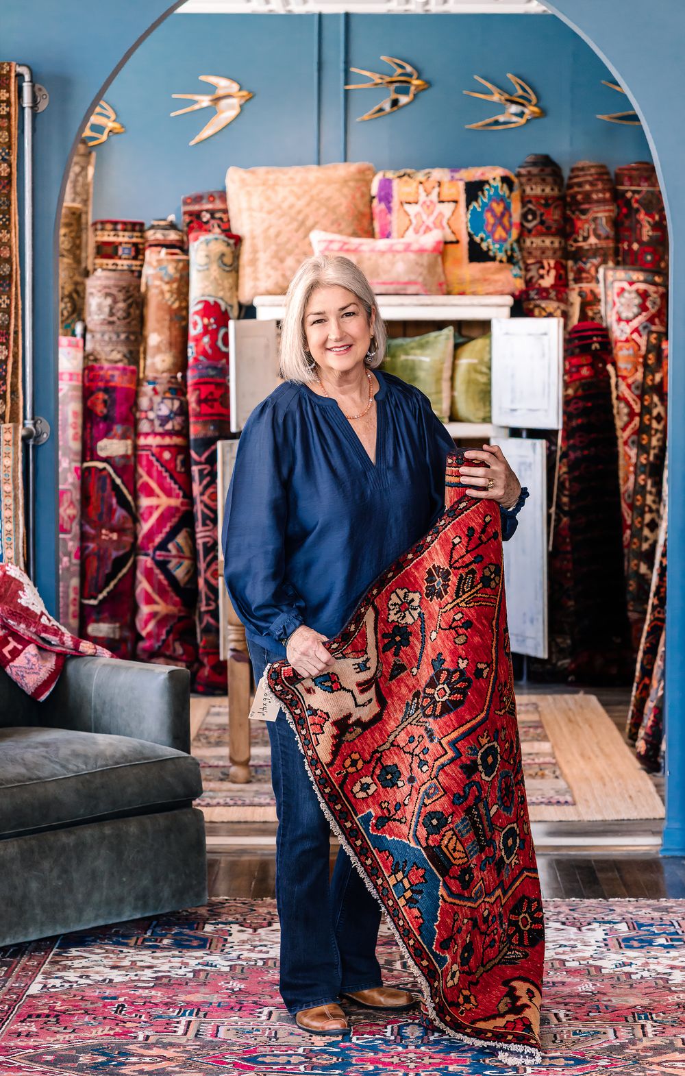 Woman in navy jumpsuit holding a colorful red Persian rug in a boutique shop filled with vibrant vintage rugs.
