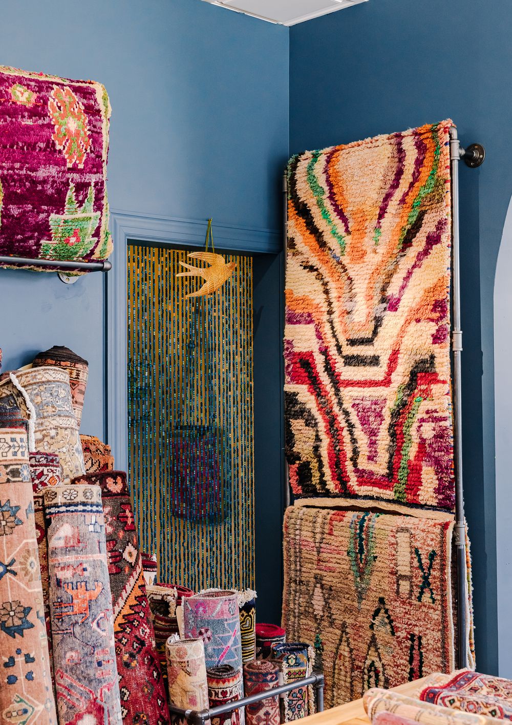 Colorful Moroccan rugs displayed on blue walls in a bohemian shop with beaded curtain doorway.