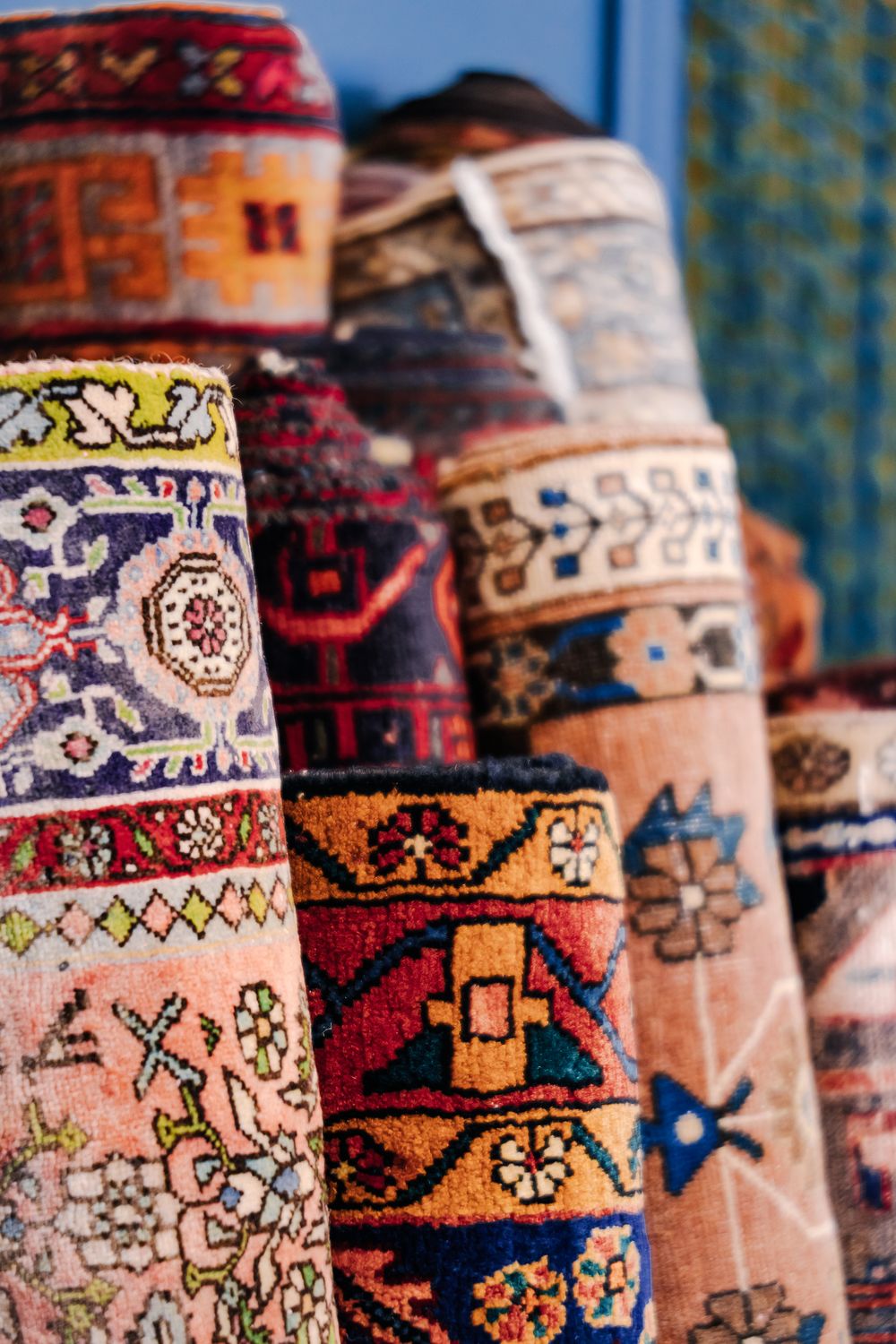 Colorful rolled Persian rugs with intricate floral patterns displayed against a blue wall in a market.