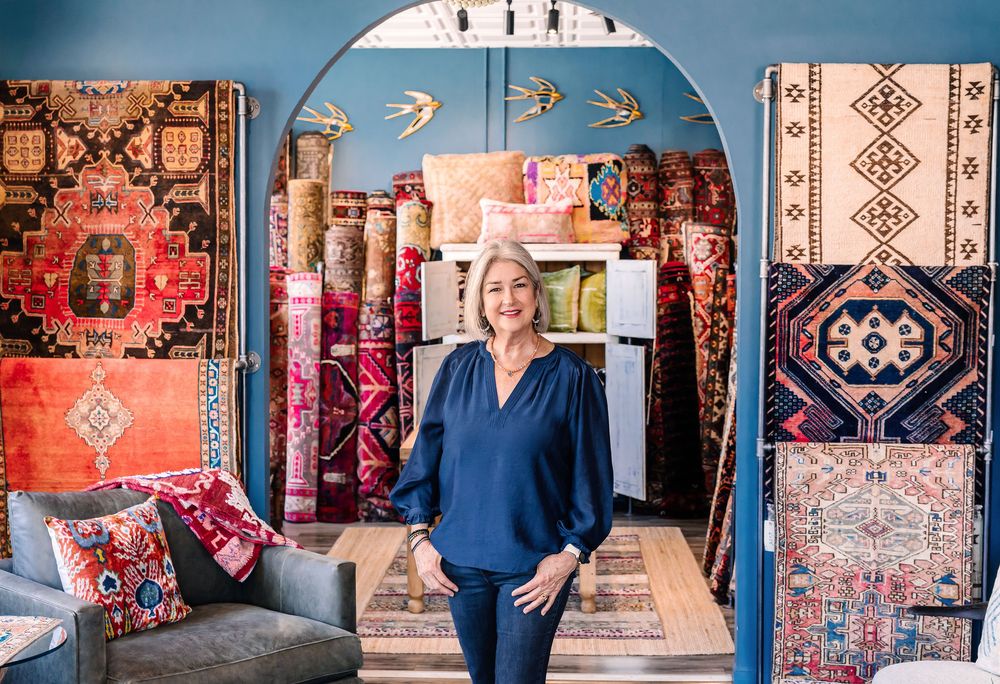 Woman stands in colorful rug shop surrounded by vibrant Persian and Turkish rugs displayed on walls and floor.