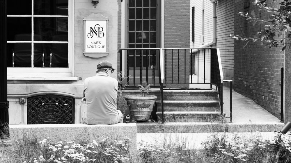 Black and white photo of a person sitting on steps outside a brick building with an NB sign.