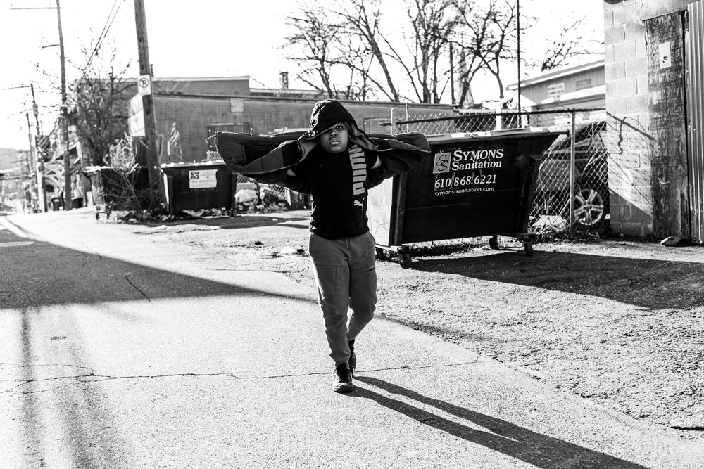 Person walking down a street carrying items over their shoulder near a dumpster in a black and white urban photo.
