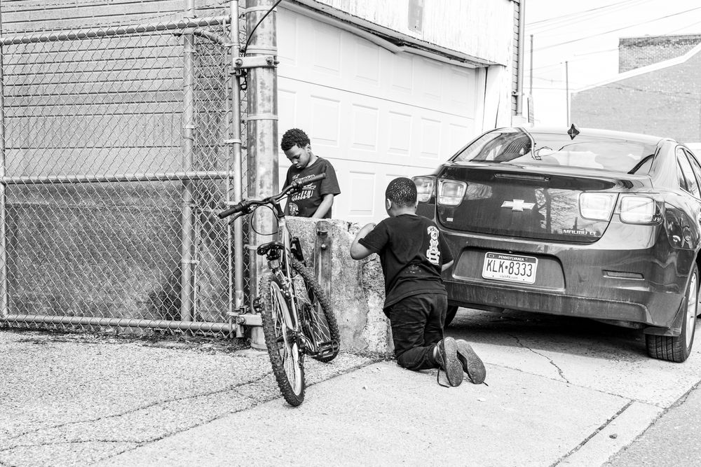 Two young men work on a bicycle near a parked car in an alley in black and white urban photo.